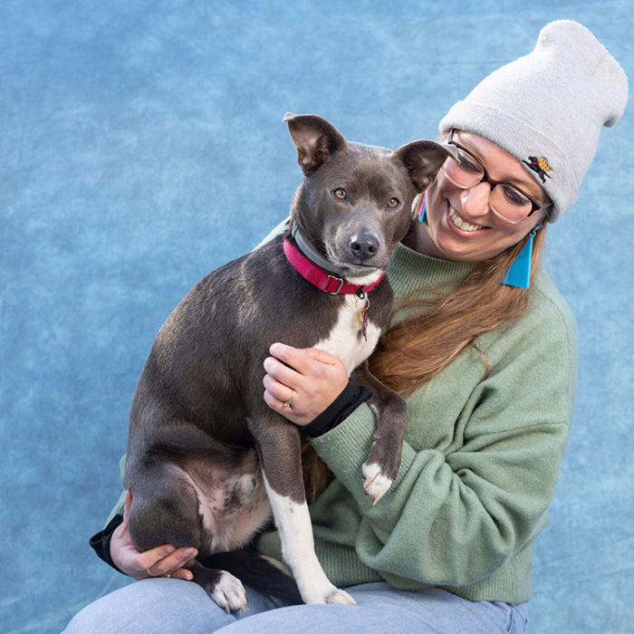 A woman wearing a gray beanie hat and a light green sweater holding her mixed breed ashy grey dog in front of a blue backdrop