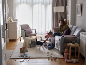 A little girl on the floor of her living room with a cat who has just come out of a cat carrier. A woman sits on the sofa behind her smiling.