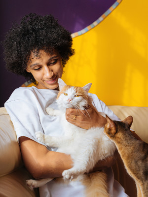 A woman with curly black hair sitting in front of a dark purple and yellow backdrop while looking down affectionately at her two cats sitting on her lap