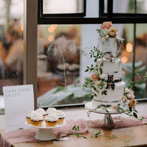 A wedding cake decorated with roses and cats sitting on a table next to a white platter of chocolate cupcakes with white icing and a sign saying "Something Sweet"