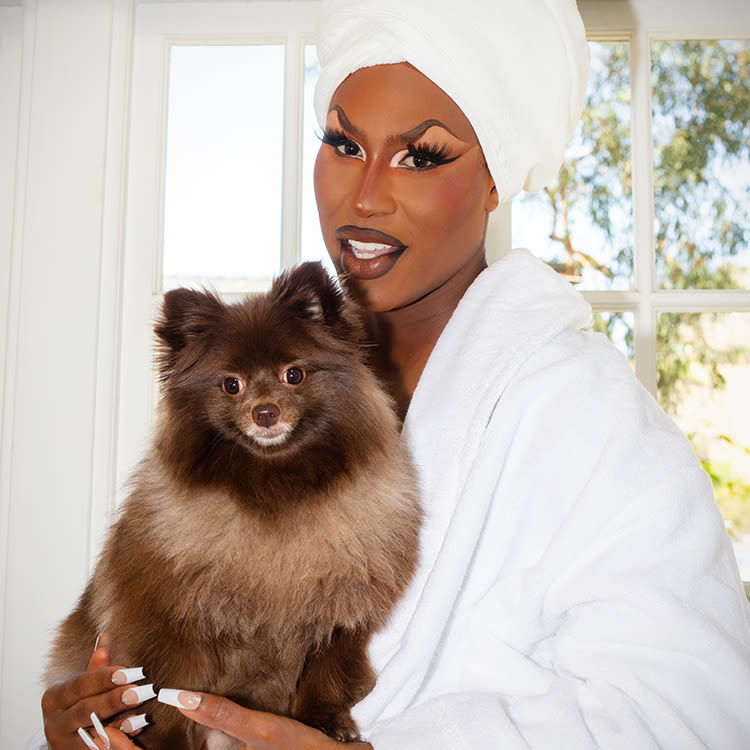 Shea Coulee in makeup and a white robe and hair towel wrap holding her Pomeranian dog Baby