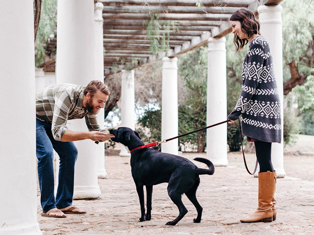 Man petting a black dog on a leash outside.