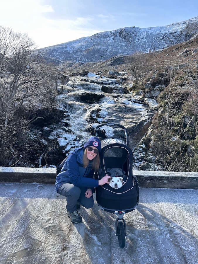 a woman crouches in front of a frozen stream with an elderly dog in a pushchair