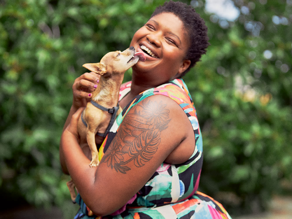 African American woman with floral arm tattoos and short curly hair laughing with joy while holding her small Chihuahua dog outside