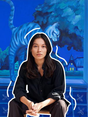 A portrait of the artist Bambou Gili, a woman with long dark hair wearing a black dress shirt and dark brown trousers sitting with one of her paintings of a cat in all blue behind her and white outlines around her figure