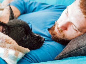 man with ginger hair and beard, wearing a blue T-shirt lying down and looking at his black dog in his arms