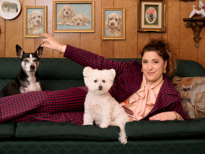 Jess Rona laying on a green couch in a vintage style living room, playfully pinching the ear of a dog sitting beside her while another white dog sitting in front of her