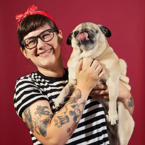 Gemma Correll smiling and holding up her one-eyed Pug dog against a red backdrop