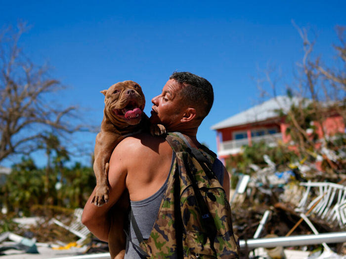 a person holds a large dog surrounded by hurricane damage