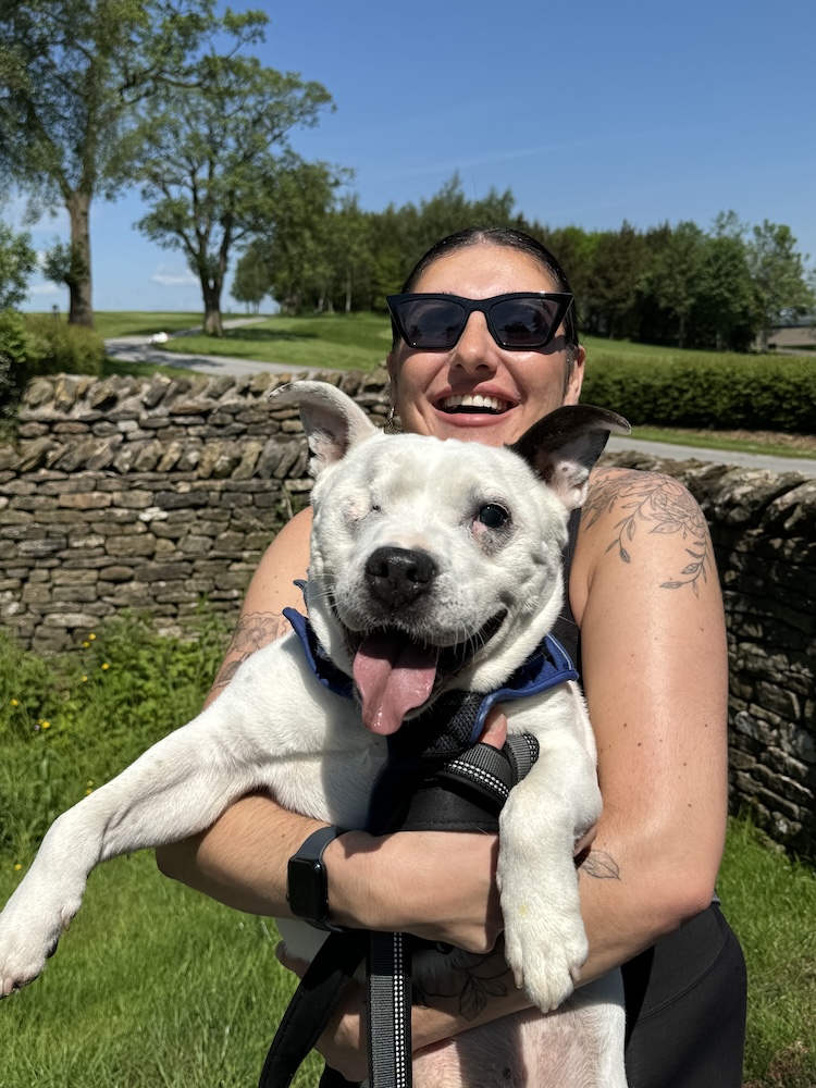 a woman in sunglasses hugs a smiling white dog with one eye