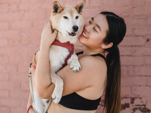 Woman with long straight black hair and a gold nose ring holding a senior Shiba Inu mixed breed dog wearing a red harness