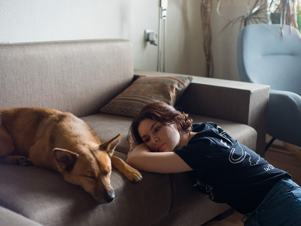 A tan dog sleeping on the couch while a short haired woman rests on her arms on the edge of the couch looking at the dog sadly