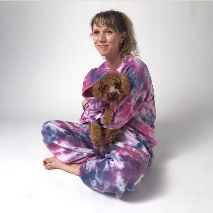A blonde woman sitting cross-legged in front of a white backdrop wearing tie-dye pyjamas with a Cavpoo dog on her lap wearing a matching tie-dye hoodie. 