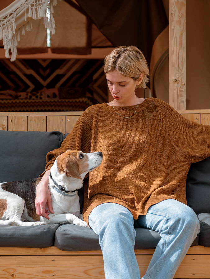Short-haired blonde woman in a tan sweater and blue jeans sitting on a bench talking to her Beagle mix dog who is looking up at her
