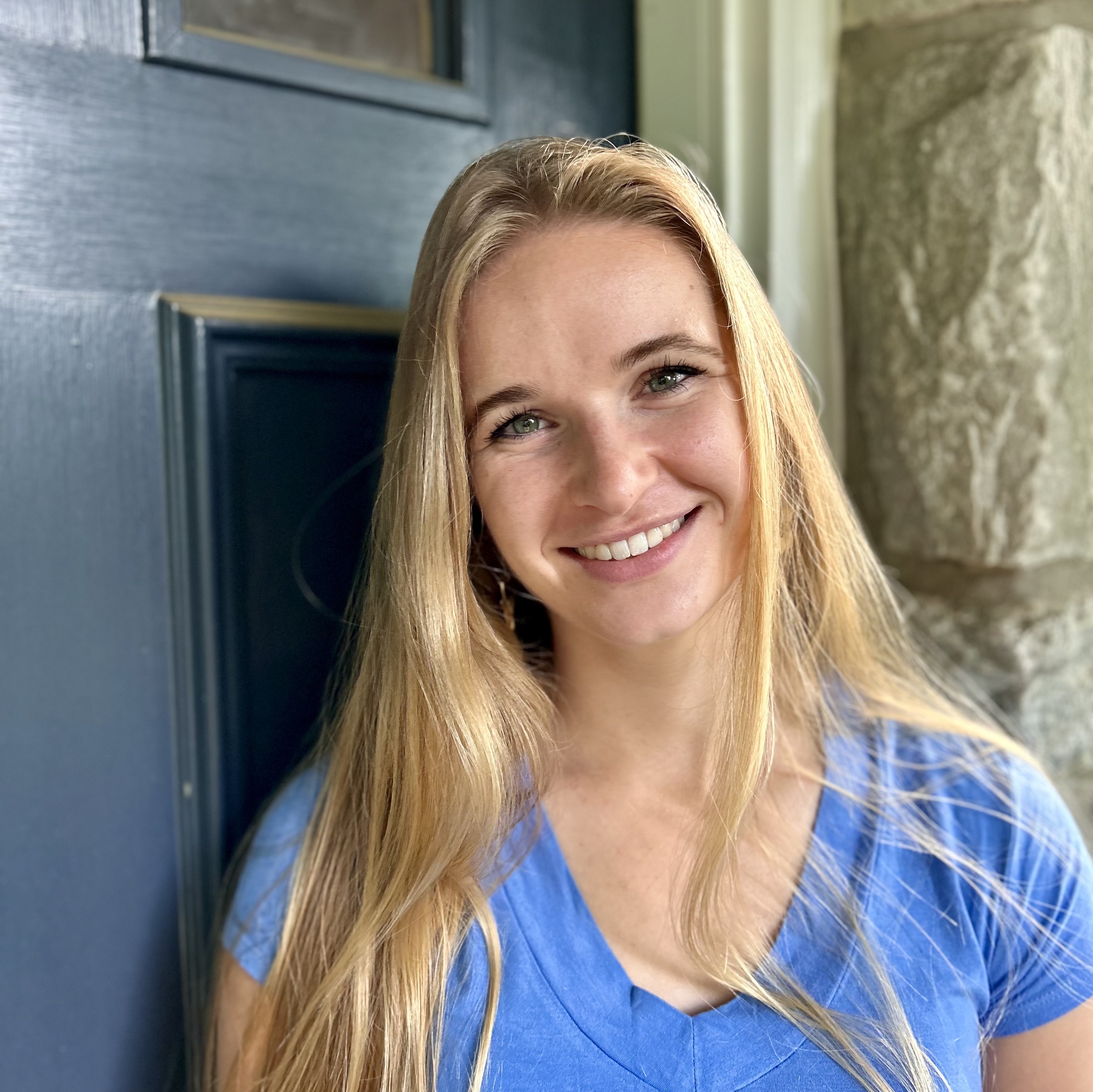 Informal headshot of a young woman white woman with long hair smiling and leaning against a blue door.}}