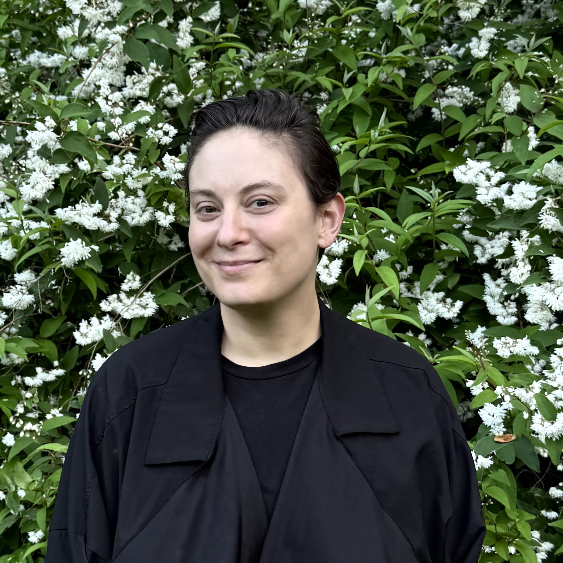 Headshot of a young person with short hair. They wear black and stand in front of a flowering bush.}}