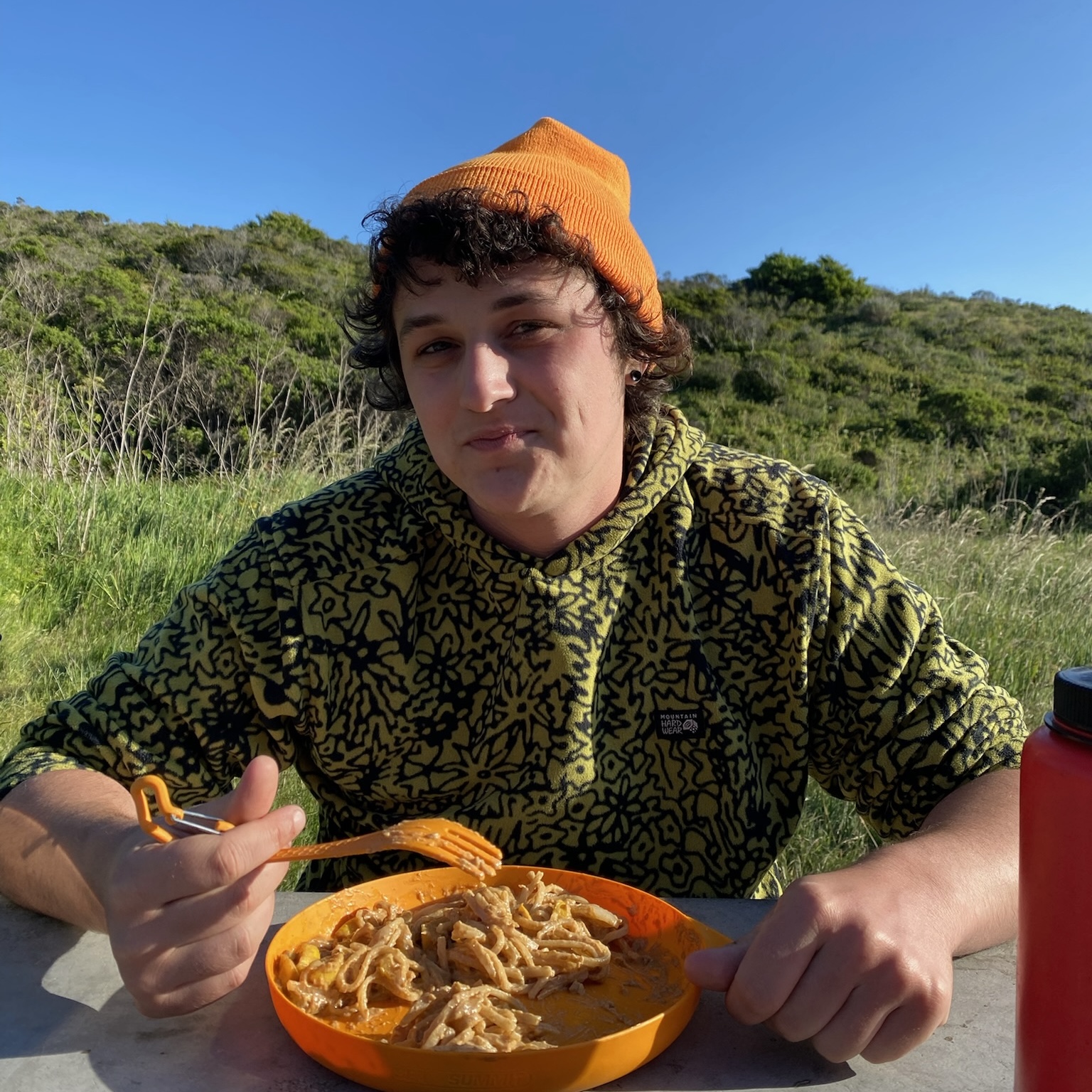 Casual photo of a young white person wearing an orange hat and eating a meal outdoors. }}