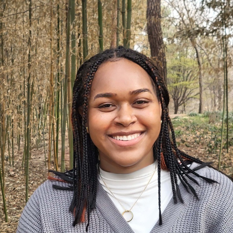 Headshot of a young Black woman with braids against a forest background.}}