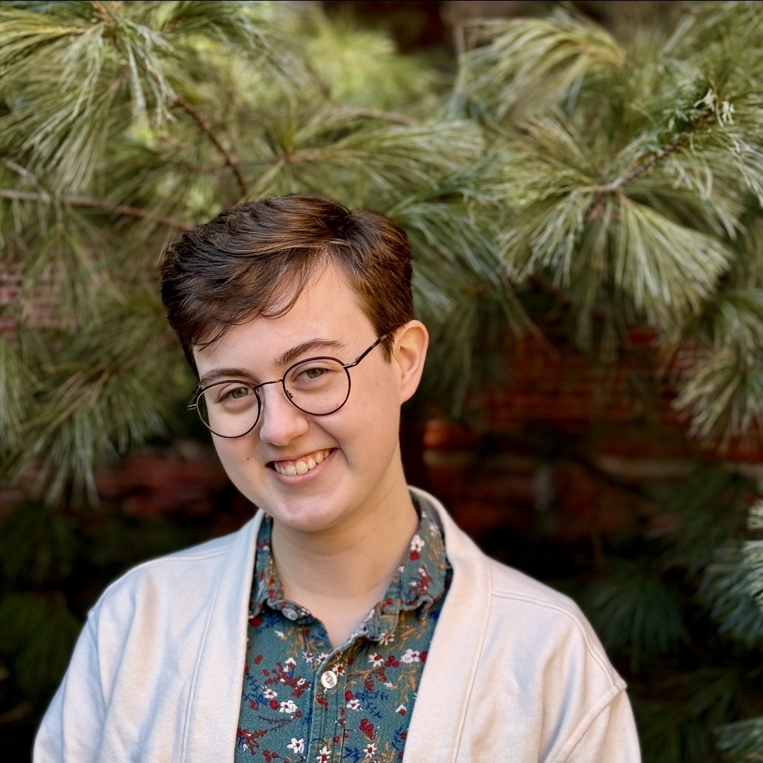 Smiling young adult with short brown hair wearing round glasses, a blue shirt, and a white cardigan.}}