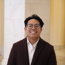 Headshot of a young man with glasses wearing a white shirt and dark blazer standing in front of a stone wall.}}