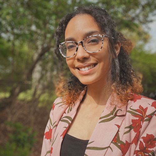 Informal headshot of a young woman with glasses and a floral blazer with trees in the background.}}