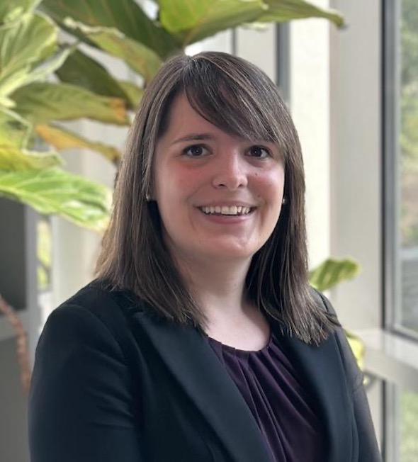 professional headshot of a young woman with brunette hair, brown eyes, a dark purple shirt and a black blazer, smiling openly in front of a large plant and a window}}