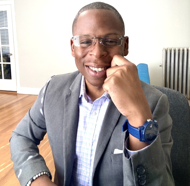 black man in a blazer seated casually in an office chair smiling at the camera}}