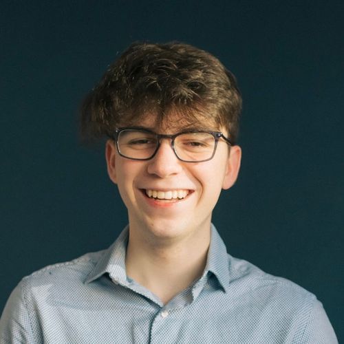 Headshot of a young man with curly hair and glasses smiling broadly at the camera.}}