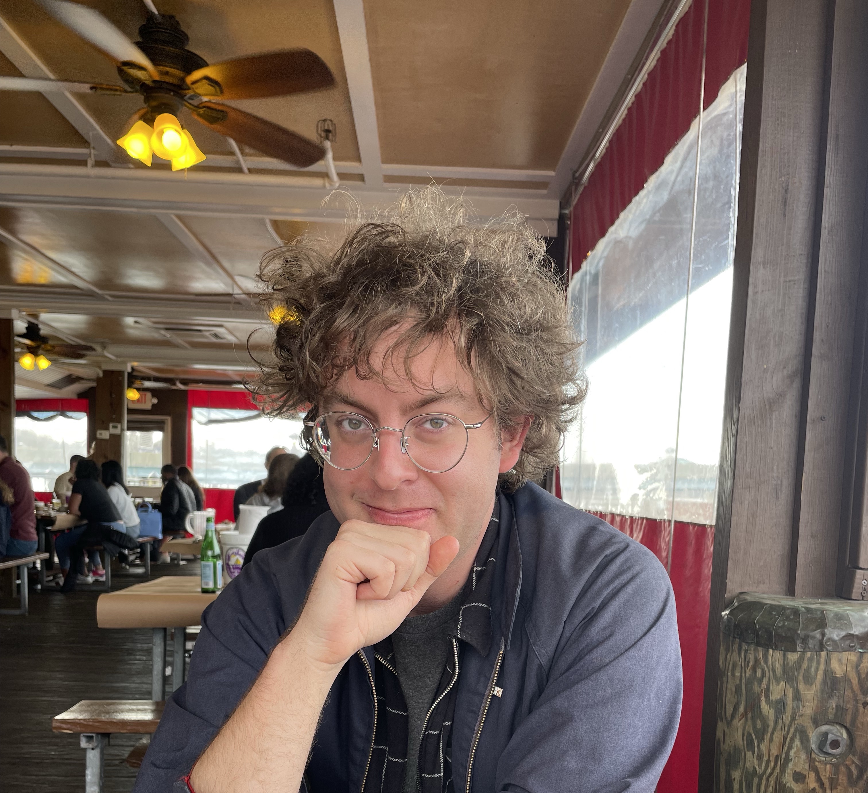 Photograph of Sam from the shoulders up. He wears glasses and a blue jacket and is holding a fist up to his smiling mouth. The interior of a Maryland crab shack is visible in the background.}}