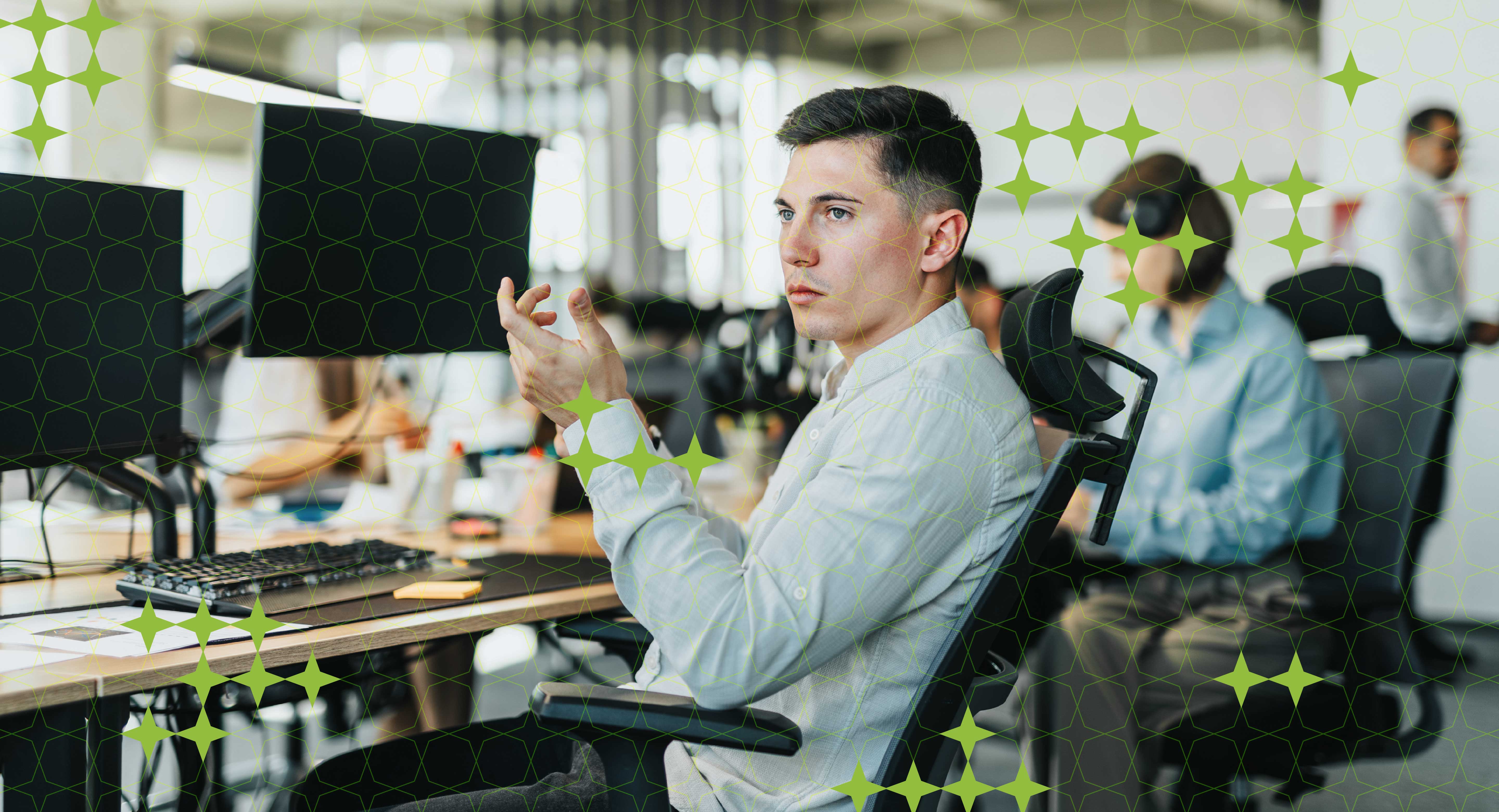 A man sits at a desk with with two monitors in an office, gesturing to a colleague not seen.