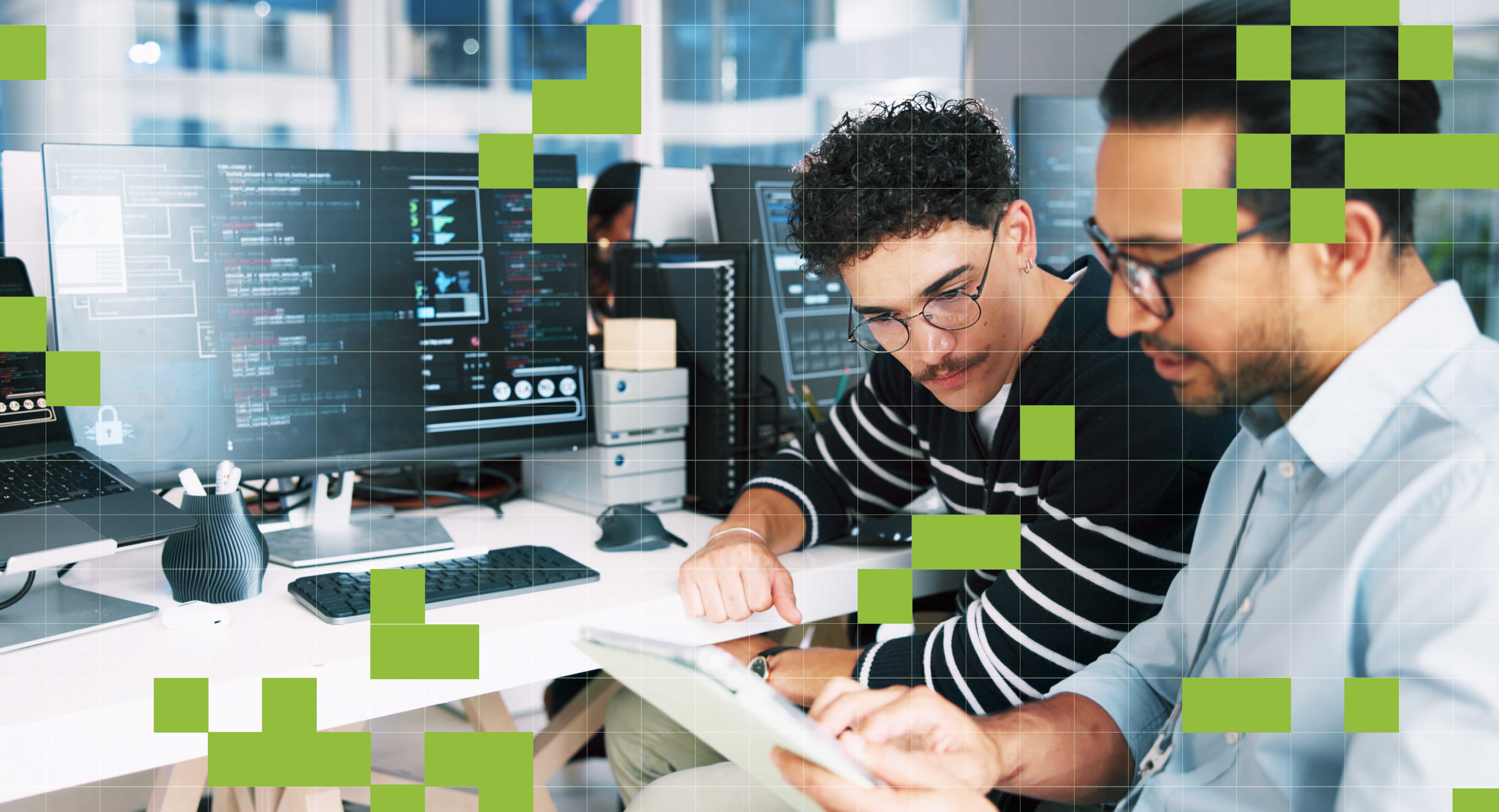 Two colleagues review items on a tablet while sitting in front of a work desktop setup.