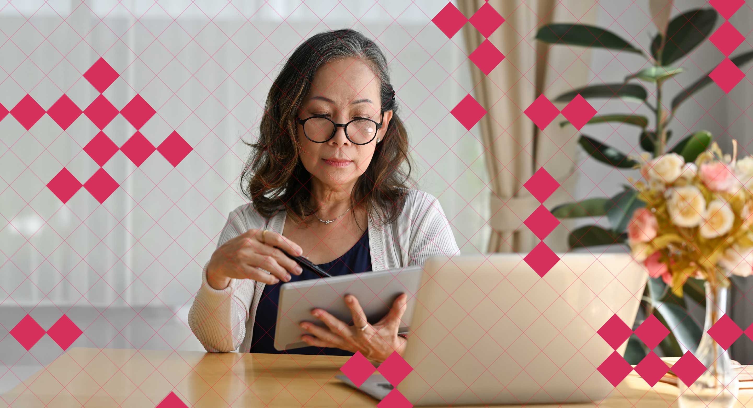 A woman sits at a desk using a tablet with a laptop on the table in front of her.