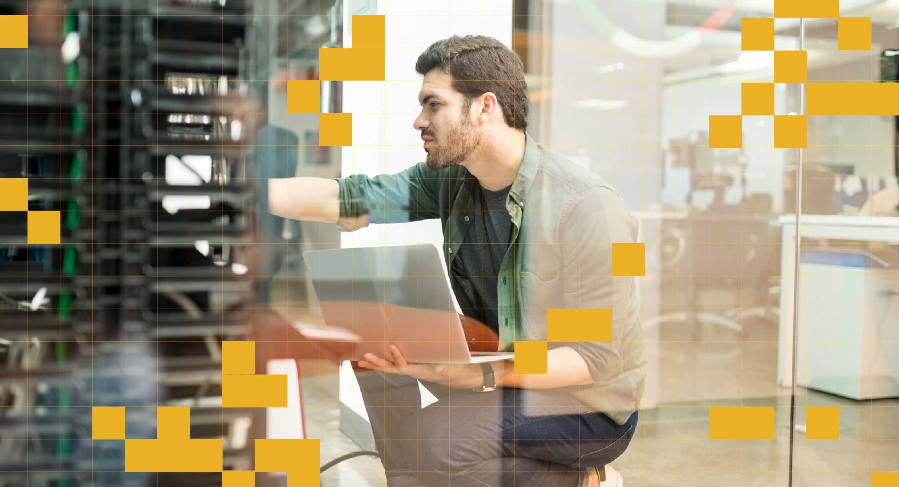 A man squats in front of a server while holding a laptop