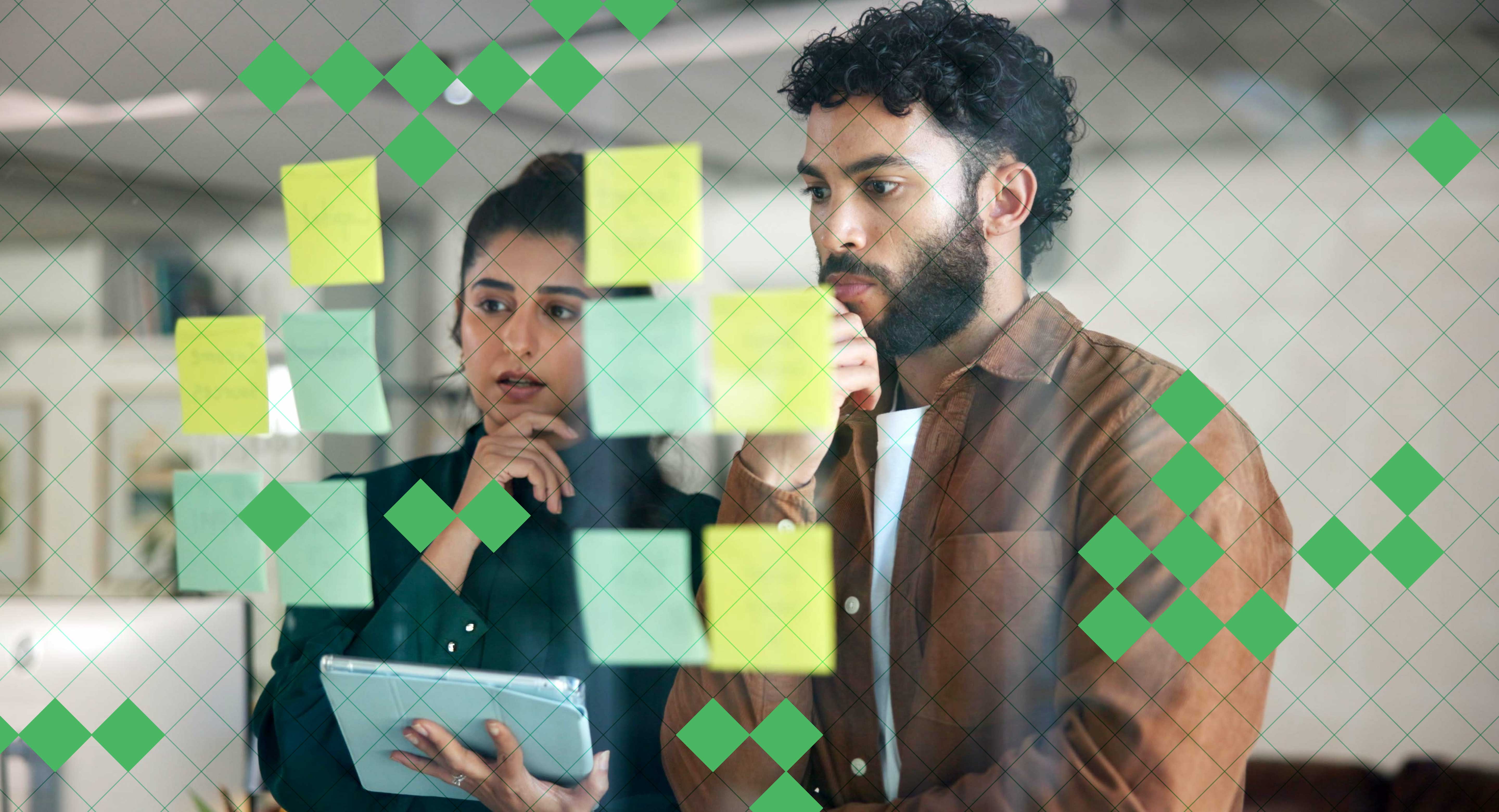Two colleagues contemplate sticky notes on a board. One is holding a tablet.