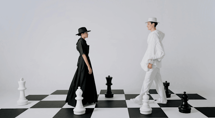 Man dressed in white and woman dressed in black standing facing each other on a giant chess board