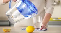 Woman pouring a glass of water from a jug with a whole lemon and lemon slices sitting on the bench
