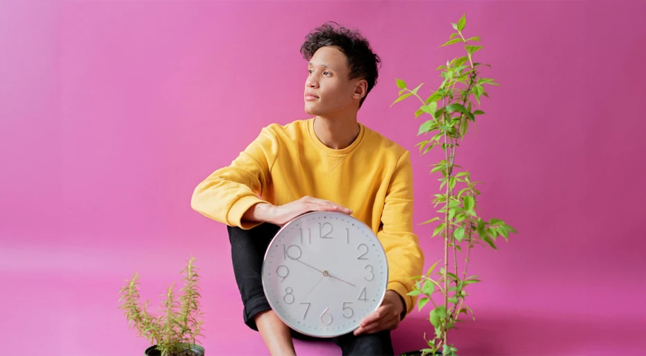 Young man sitting on the ground against a pink background, holding a wall clock in his hands. 