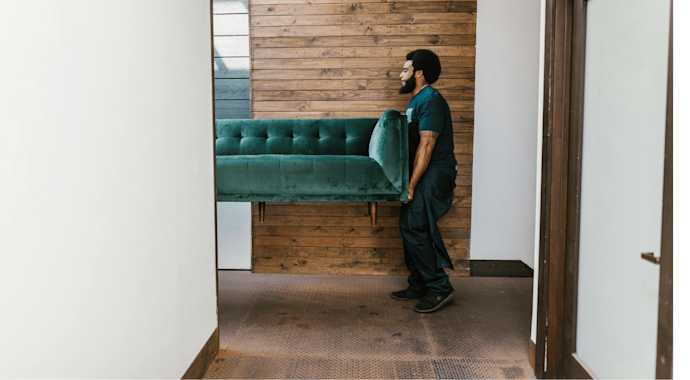 Man carrying one end of a teal green couch, with the other person hidden behind a wall.