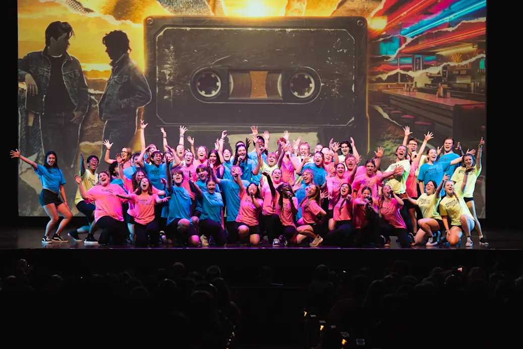 A large group of performers in pink and blue shirts pose on a stage with arms raised