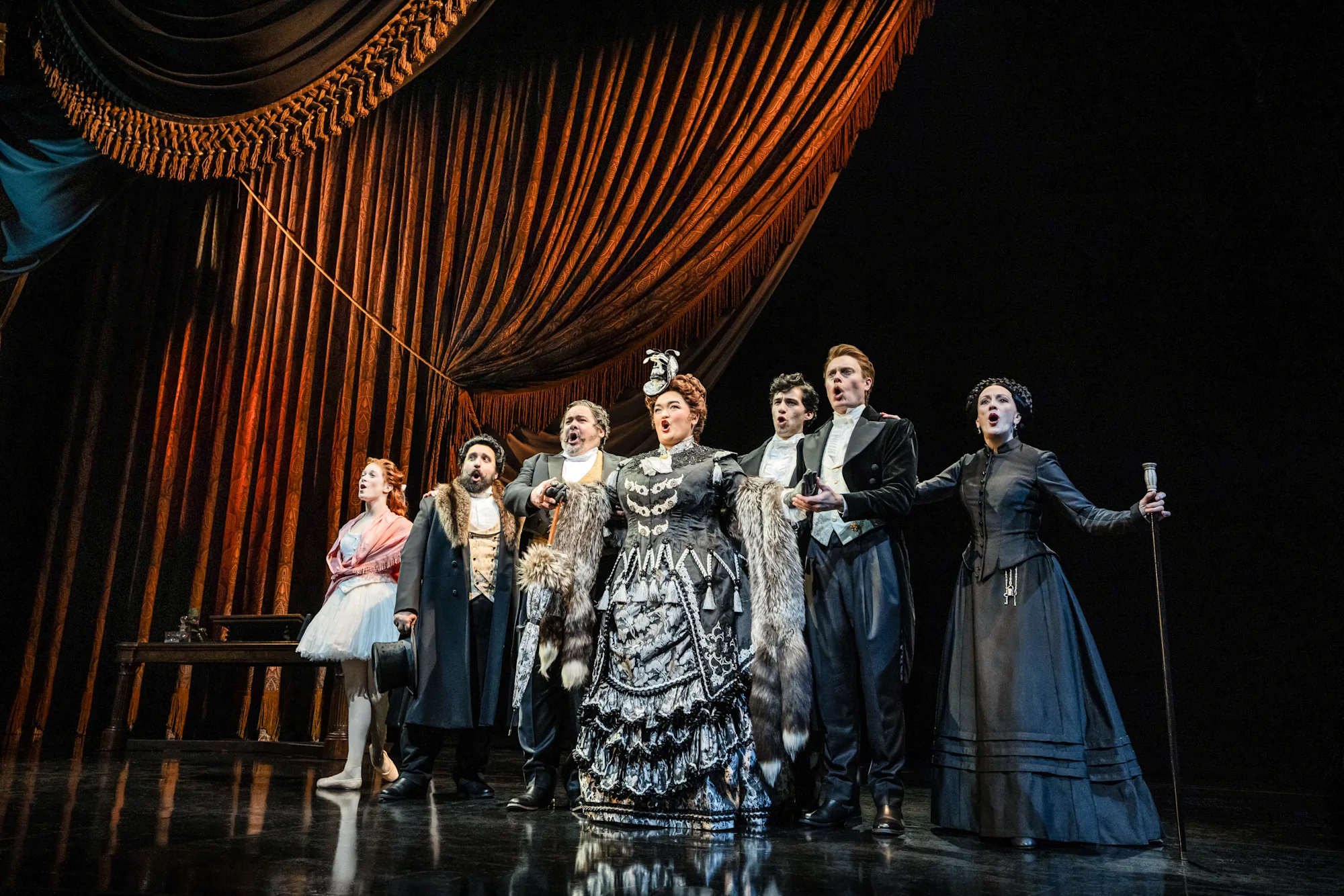 "Prima Donna" from The Phantom of the Opera North American Tour. (L to R) Melo Ludwig, Christopher Bozeka, William Thomas Evans, Midori Marsh, Daniel Lopez, Carrington Vilmont, Lisa Vroman; Photo: Matthew Murphy and Evan Zimmerman; The cast bellows out a collective tune with a burgundy curtain held open with one rope and poised to close;