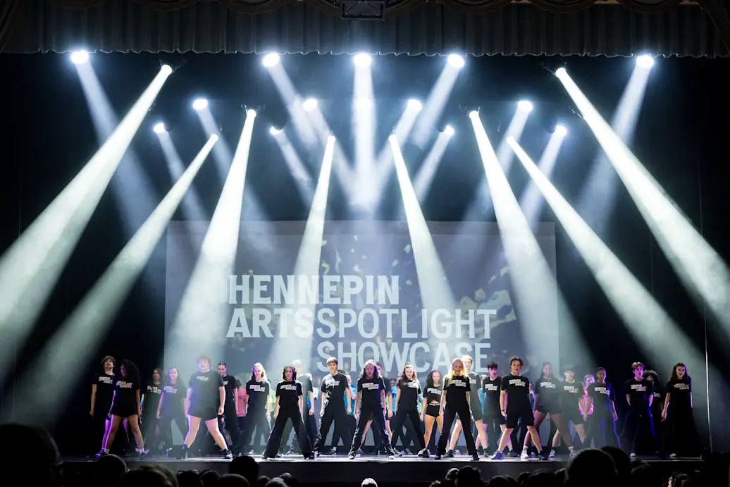 Performers on a stake in staggered rows stand still with beam of light from cascading down around them and Hennepin Arts Spotlight Showcase behind them