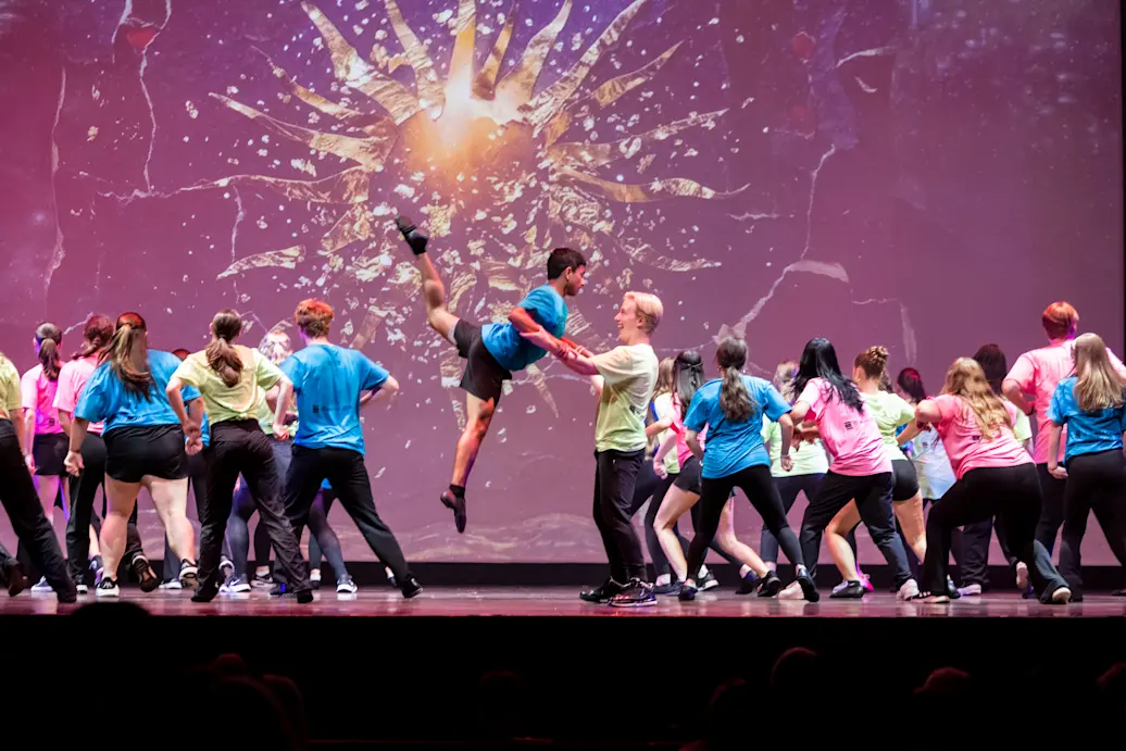 Dancers in bright shirts perform on a stage with two central dancers doing a leap. Credit: Dan Norman