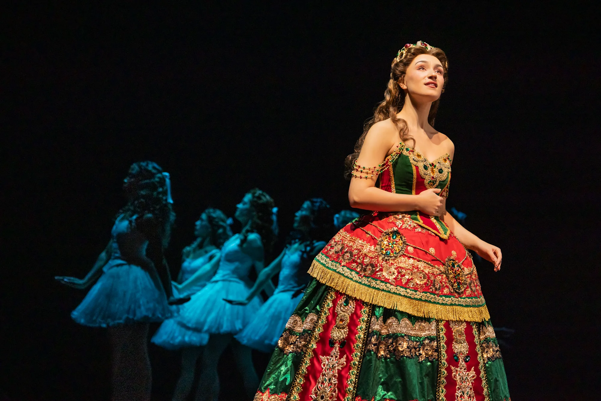 Jordan Lee Gilbert as Christine Daaé in The Phantom of the Opera North American Tour; Photo: Matthew Murphy and Evan Zimmerman; Christine wears an ornate red and green dress and looks wistfully upward with hand on abdomen with ghost-like dancers in the background;