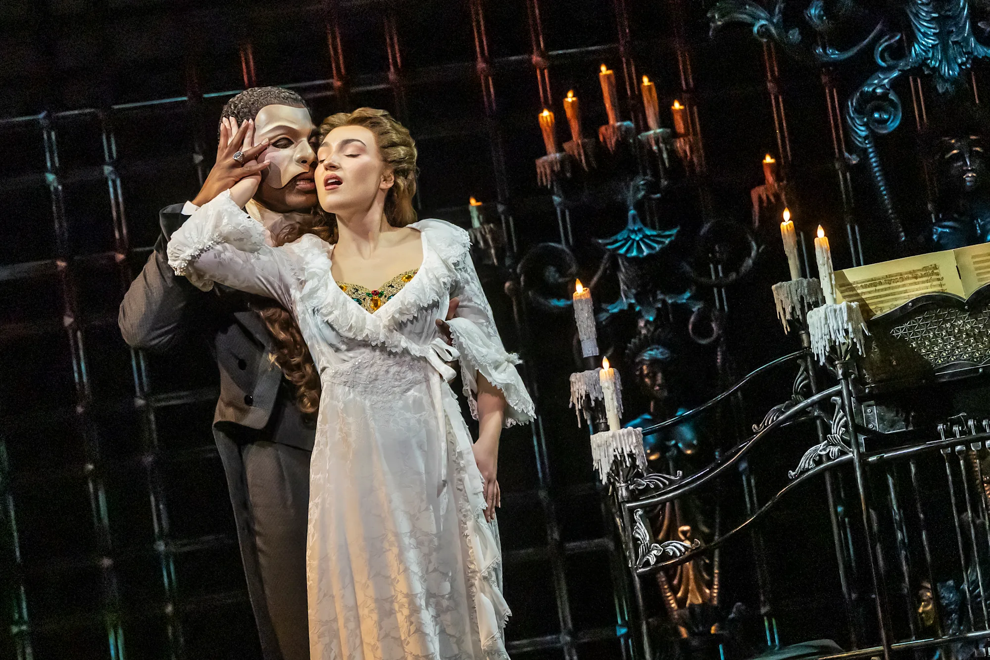 (L to R) Isaiah Bailey as The Phantom and Jordan Lee Gilbert as Christine Daaé in The Phantom of the Opera North American Tour; Photo: Matthew Murphy and Evan Zimmerman; Christine leans back and puts her hand onto the Phantom's masked face, with lots of candles around;