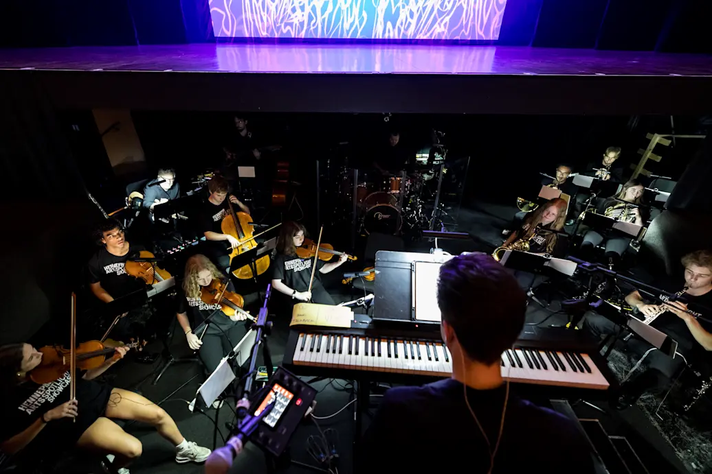 View of an orchestra below stage level with a keyboard in the foreground, violins and woodwinds on the side, and a stage in top of the photo with a colorful background