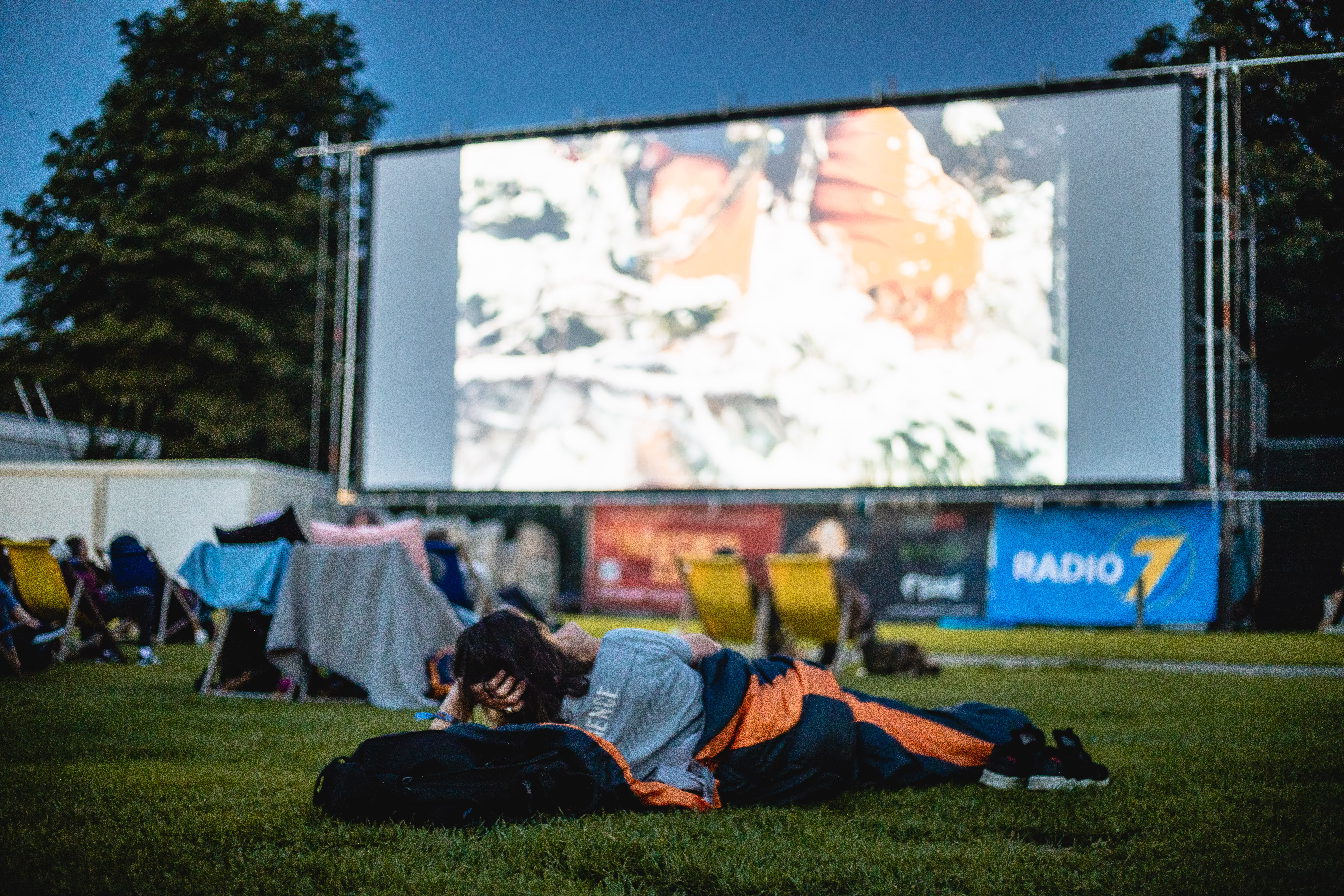 Eine Person liegt im Schlafsack auf der Grünfläche vor der Leinwand im Open Air Kino