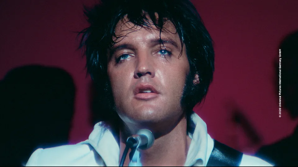 Foto aus einer bisher unveröffentlichten Konzertaufnahme von Elvis Presley. Er steht vor dunkelrotem Hintergrund auf der Bühne und trägt ein weißes Hemd.