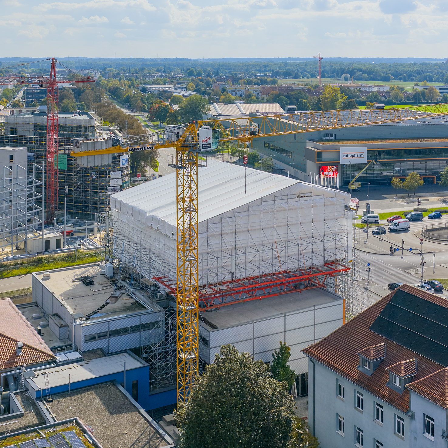 Luftaufnahme des Dietrich Theater Kino 5, das vollständig eingerüstet und mit weißer Schutzplane umhüllt ist. Daneben ragt ein gelber Baukran empor. Im Hintergrund stehen weitere Gebäude, Baustellen und eine vielbefahrene Kreuzung.