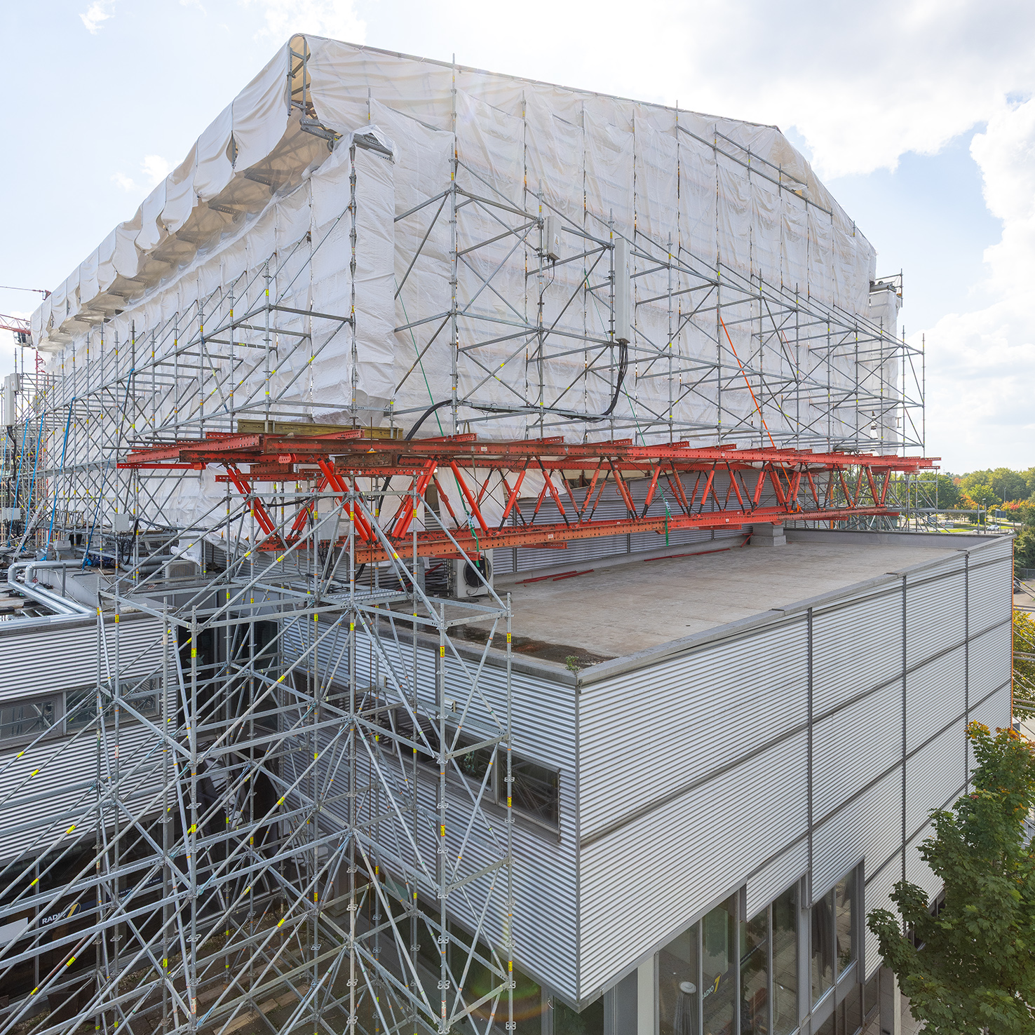 Baustellenszene des Dietrich Theater Kino 5, das dicht eingerüstet und mit weißer Schutzplane umhüllt ist. Unterhalb sind rote Tragekonstruktionen und metallene Fassadenelemente zu sehen. Rechts ragt ein Baum ins Bild.