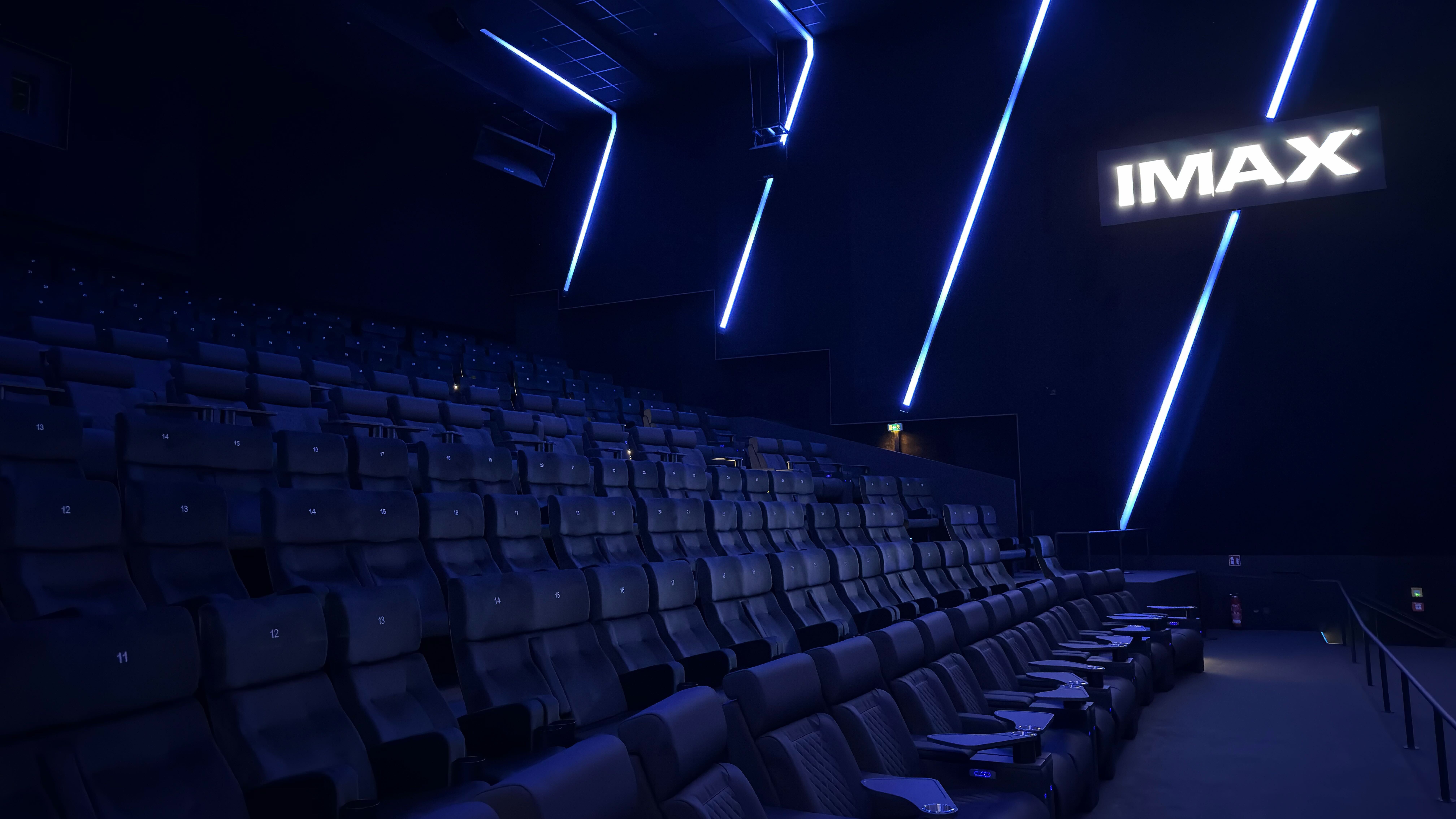 Moderner IMAX-Kinosaal mit Reihen aus dunklen, gepolsterten Sitzen, blauer LED-Lichtinszenierung an Wänden und Decke sowie leuchtendem IMAX-Logo im Hintergrund.